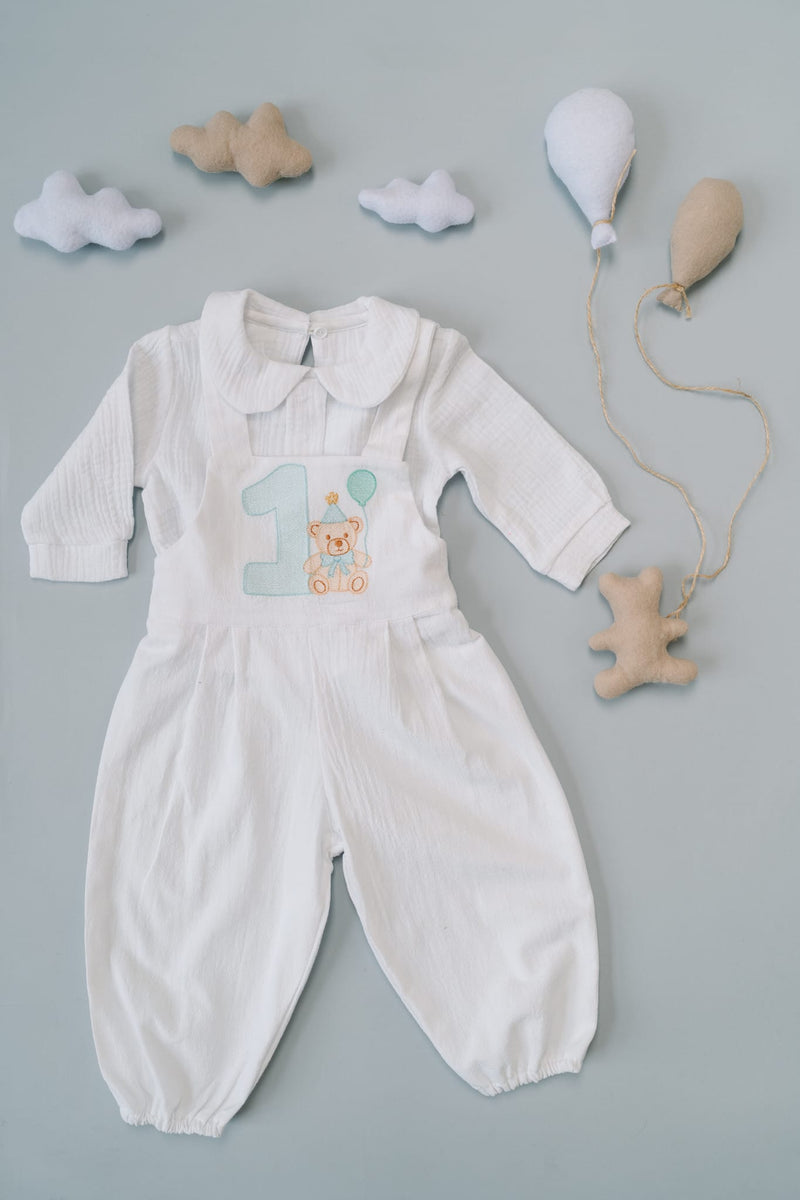 A white onesie with a number one on it, along with some stuffed animals and balloons, is displayed on a light blue background.