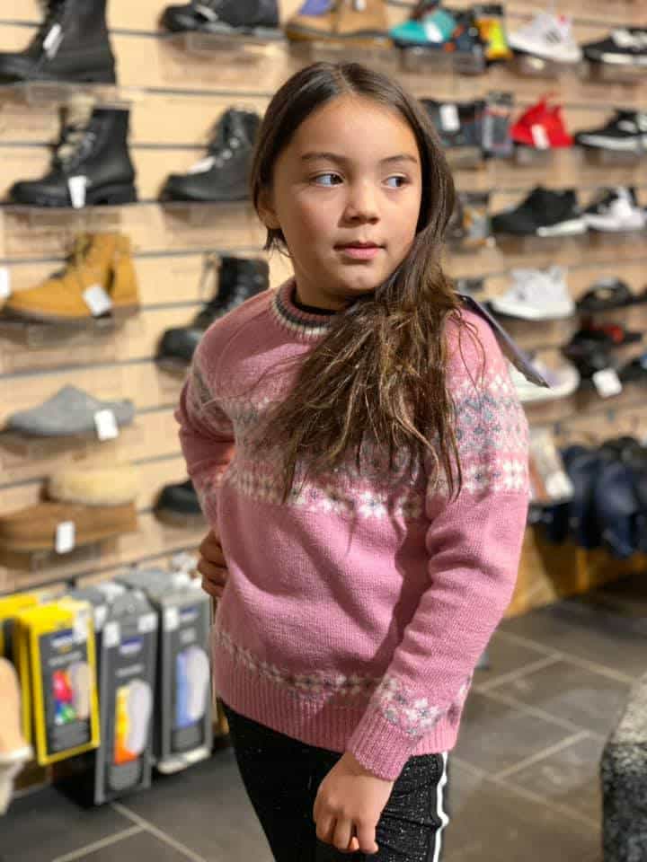 A young girl with long brown hair stands in a shoe store, wearing a pink sweater with a patterned design. She is surrounded by various pairs of shoes on display, creating a visually striking scene.