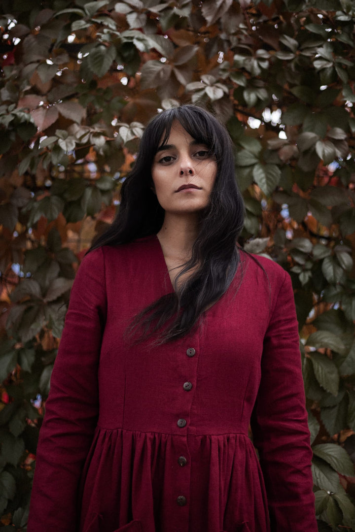 A woman in a red dress stands in front of a backdrop of autumn leaves, her gaze directed off to the side.
