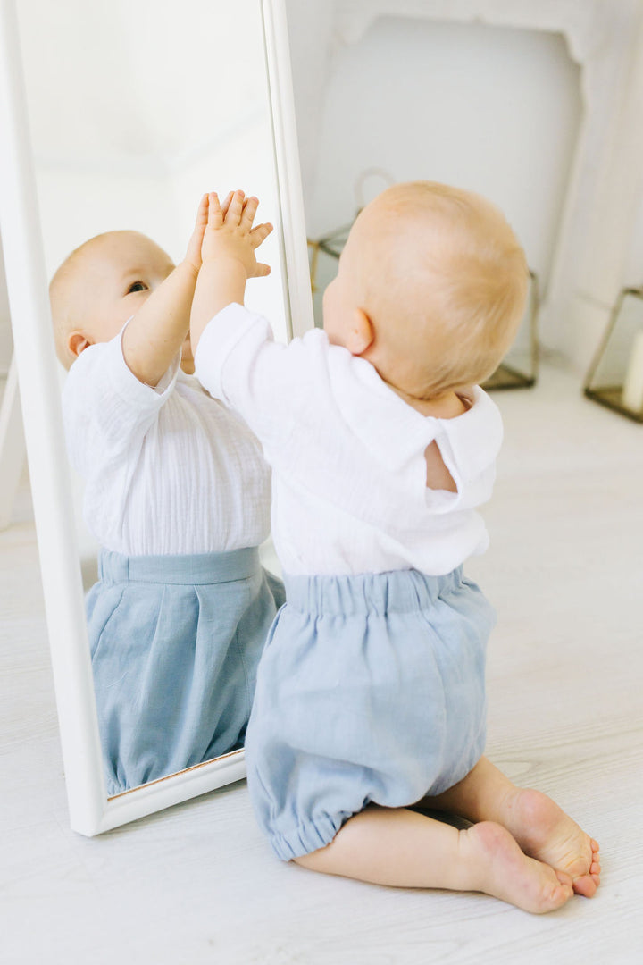 A baby in a white shirt and blue shorts is reaching up to touch their reflection in a mirror, with their hands raised.