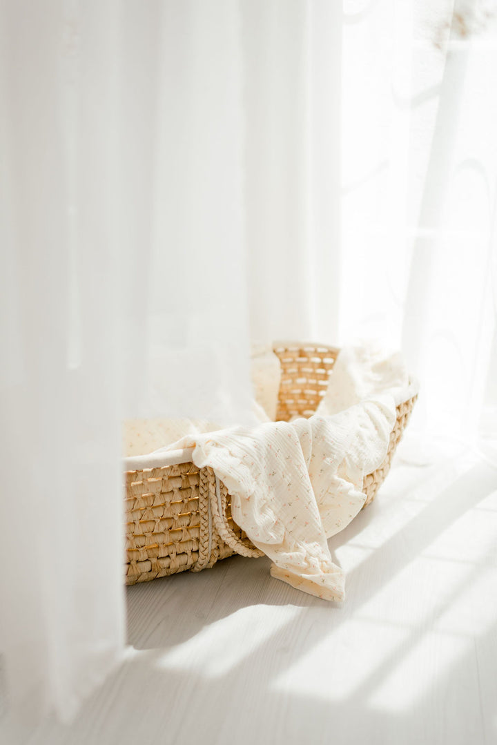 A woven basket with a white blanket draped over it sits on a wooden floor, bathed in soft, natural light filtering through sheer white curtains.