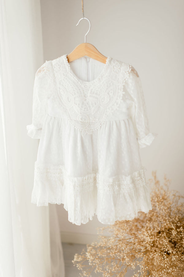 A white lace dress with a ruffled collar and sleeves is hanging on a wooden hanger against a white wall.
