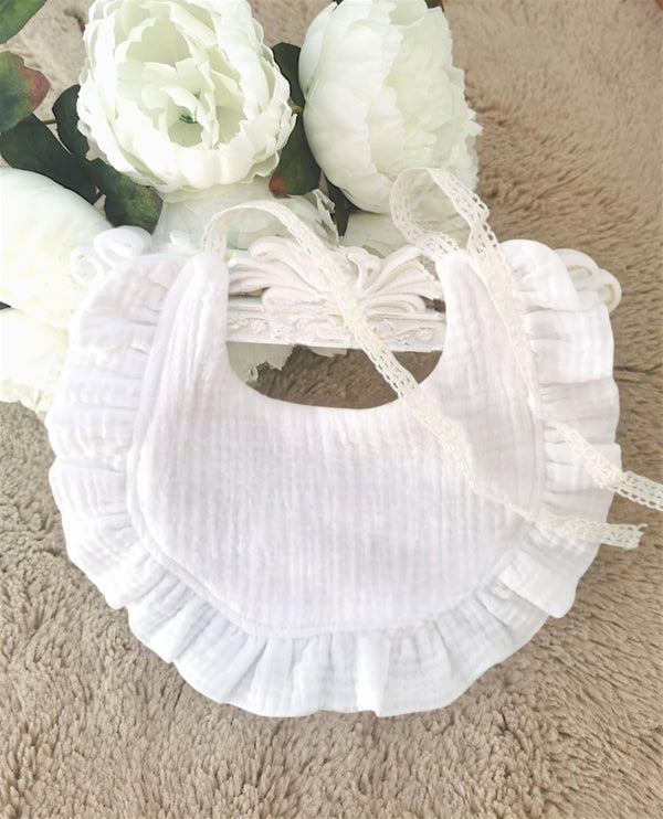 A white ruffled bib with a lace trim is placed on a beige surface, accompanied by a bouquet of white flowers.