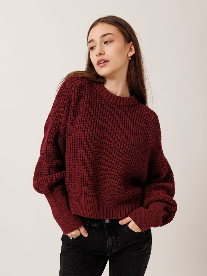 A woman wearing a burgundy knitted sweater stands against a plain background, looking to her left with a thoughtful expression.