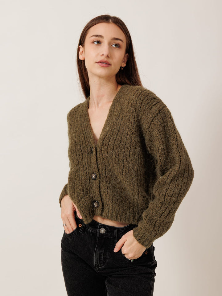 A woman wearing a green cardigan and black pants stands against a plain background, looking off to the side.