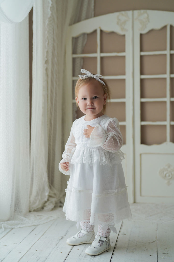 A young girl in a white lace dress and bow stands in front of a white curtain, smiling at the camera.