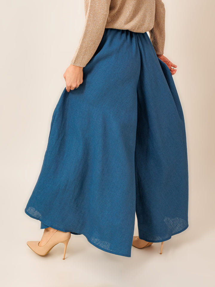 A woman wearing a long, flowing blue skirt and beige high heels is shown from behind, with her hands behind her back.