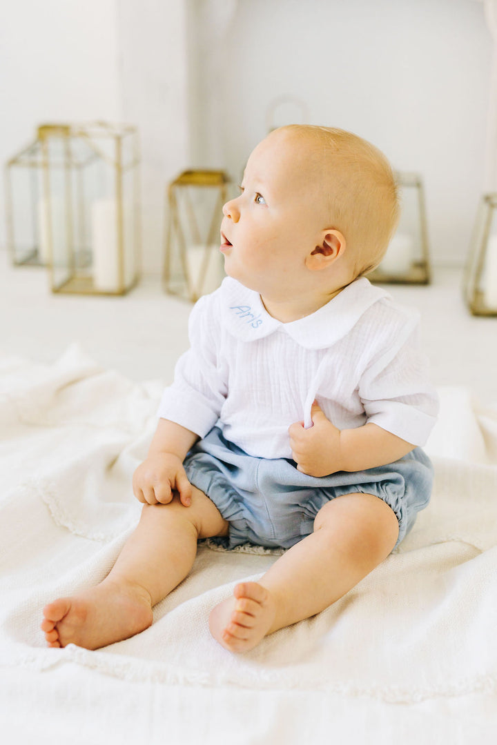 A baby sitting on a white blanket, looking up with a curious expression.