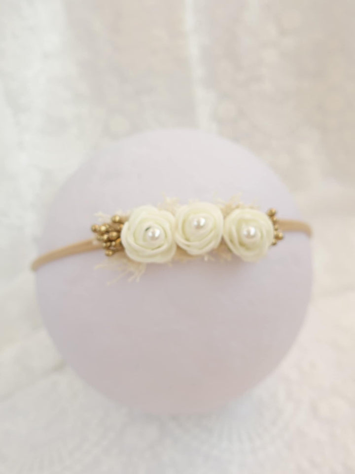 A white headband adorned with three white roses and gold beads is displayed against a white background.
