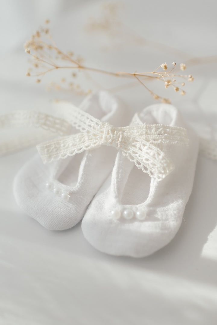 A pair of white baby shoes with a delicate lace bow, placed on a white surface.