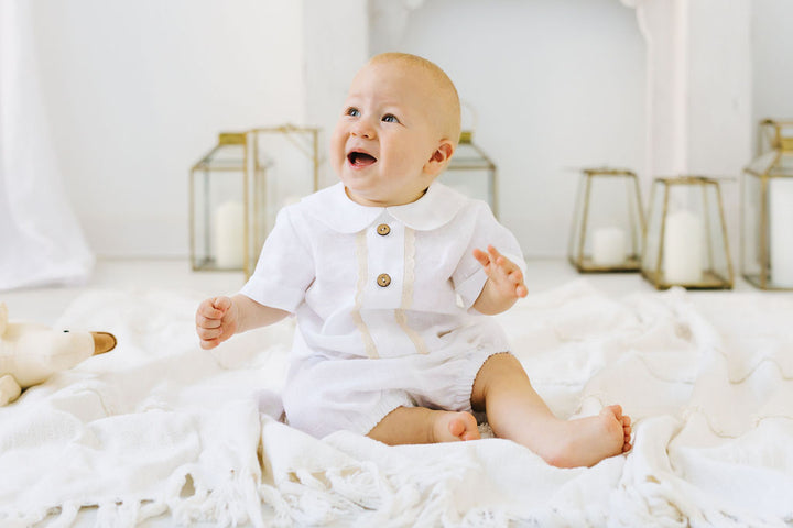 A smiling baby in a white outfit sits on a white blanket, surrounded by candles and a stuffed animal.