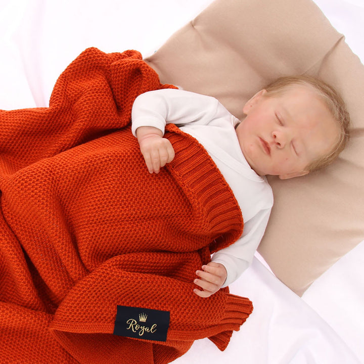 A newborn baby sleeps peacefully, wrapped in a red blanket with a black label that reads "Royal".