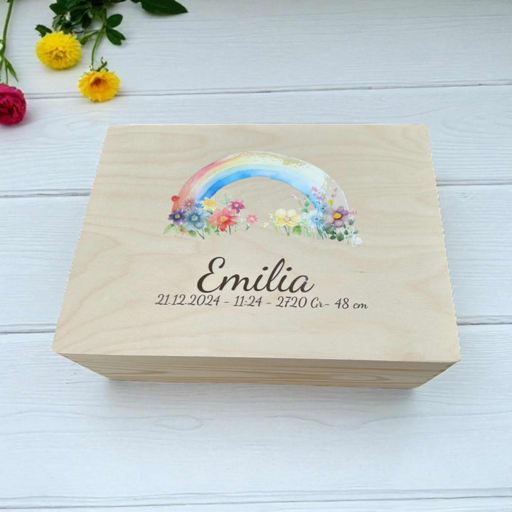 A wooden box with a colorful rainbow graphic and the name "Emilia" printed on it, placed on a white wooden surface.