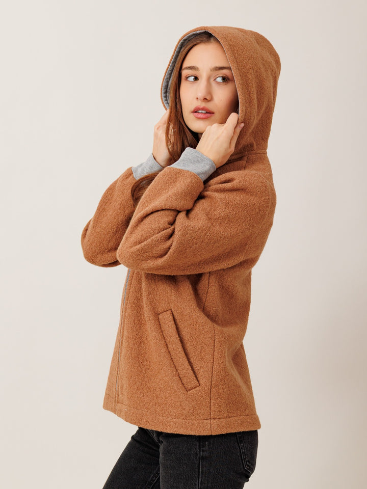 A woman wearing a brown hooded jacket stands against a plain background, looking to the side with her hands on her face.
