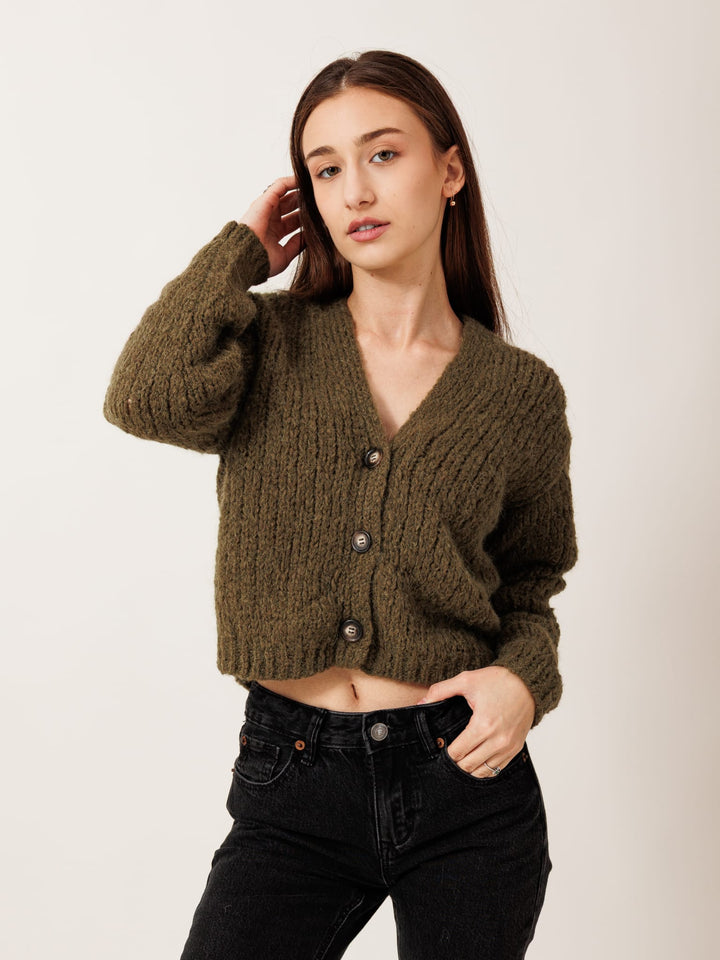 A young woman with long brown hair wearing a brown cardigan and black pants stands against a plain white background, posing with her hand on her hip.