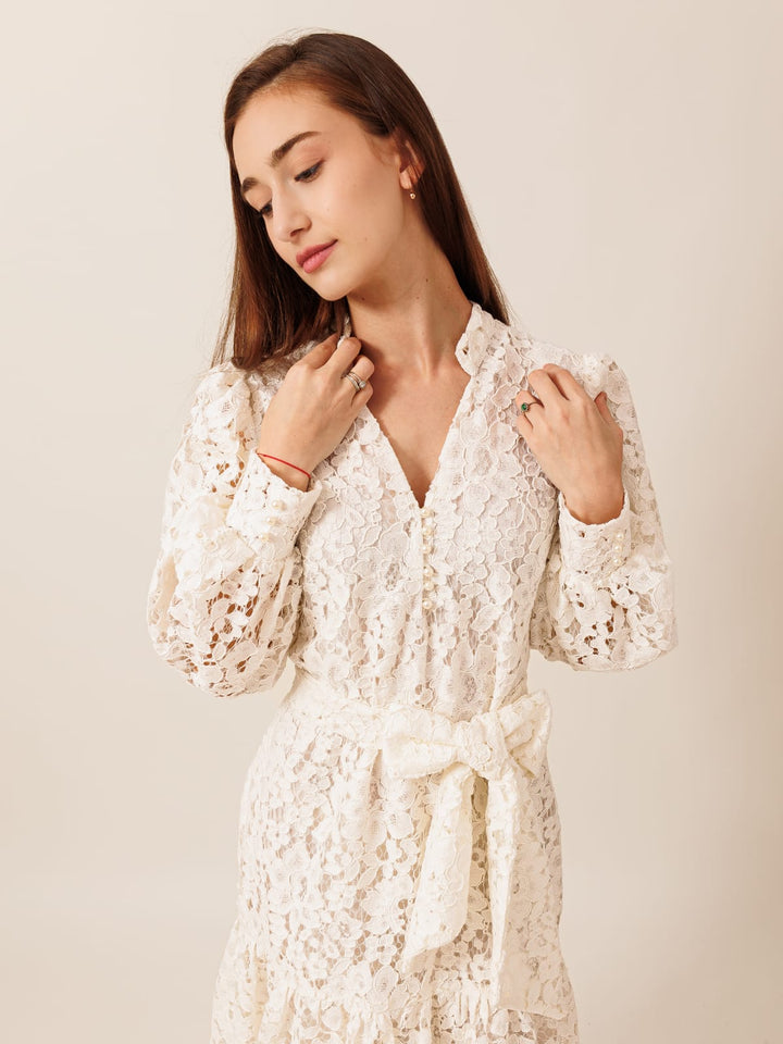 A young woman wearing a white lace dress with a bow at the waist, posing with her hands on her shoulders against a plain background.