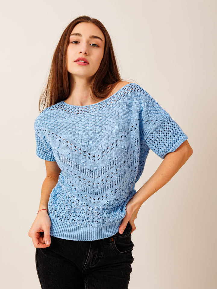 A young woman with long brown hair wearing a light blue knitted top and black pants stands with her hand on her hip, looking directly at the camera.