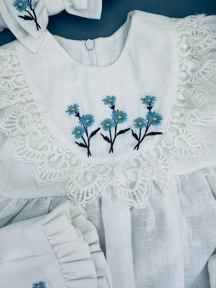 A white dress with blue flowers embroidered on the front, featuring a lace collar and ruffled sleeves.