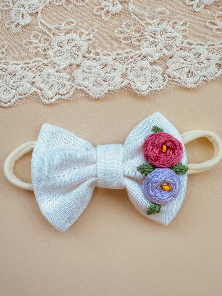 A white bow with pink and purple flowers attached to it, placed on a beige background with a lace doily.