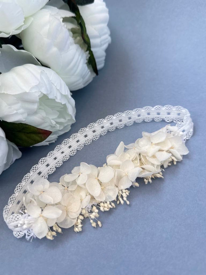 A delicate white lace headband adorned with small white flowers rests on a blue surface, accompanied by a bouquet of white flowers.