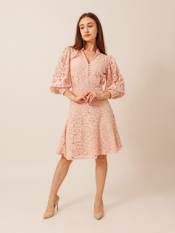 A woman wearing a pink lace dress with puffed sleeves and a short skirt, posing confidently with her hands on her hips.