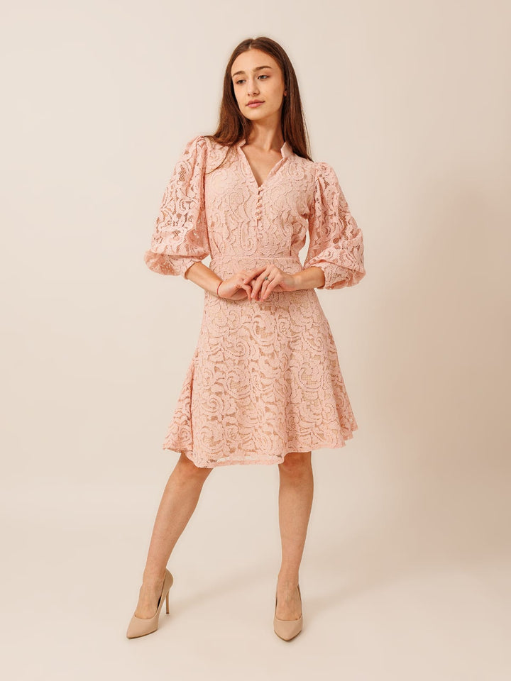 A woman wearing a pink lace dress with puffed sleeves and a short skirt, posing confidently with her hands on her hips.