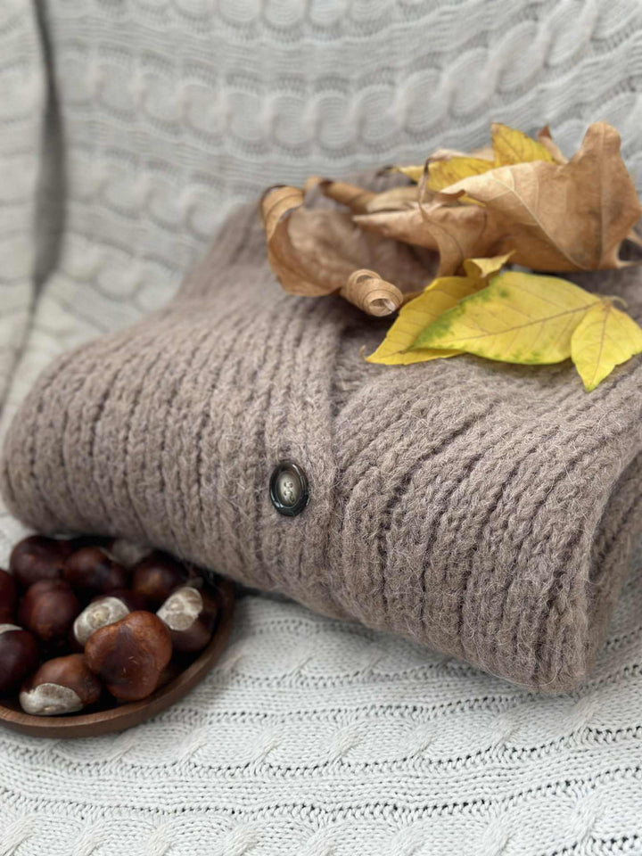 A cozy gray sweater with a button is draped over a white blanket, accompanied by a small wooden bowl filled with chestnuts and autumn leaves.