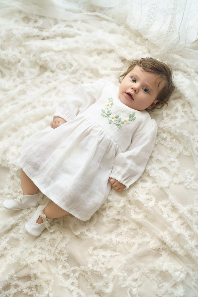 A baby girl lies on a white lace bedspread, wearing a white dress with green embroidered flowers.