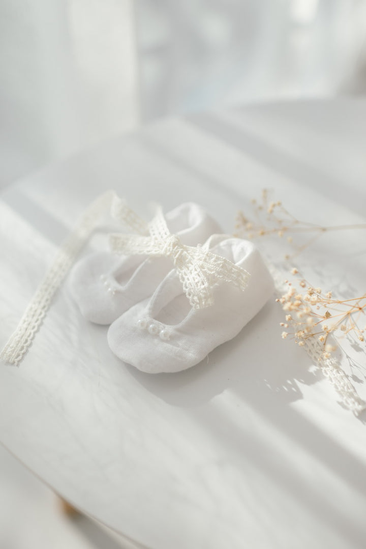 A pair of white baby shoes with a lace bow sits on a white surface, accompanied by delicate white flowers.