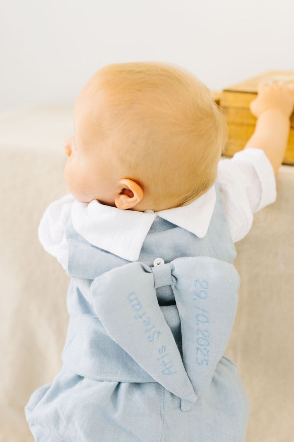 A baby wearing a light blue outfit with a bow on the back, standing against a white background.