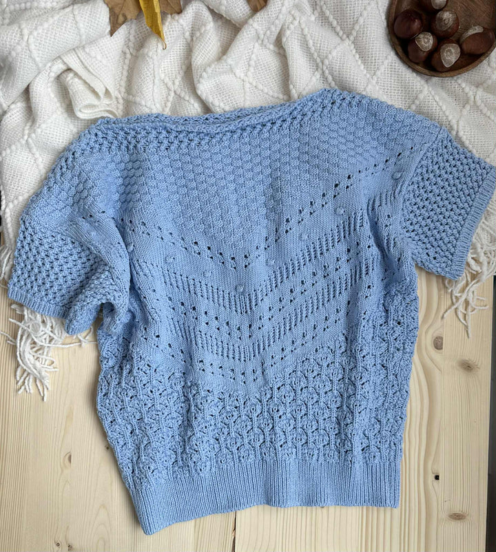 A light blue knitted sweater with a zigzag pattern is laid out on a wooden surface, accompanied by a white blanket and a bowl of nuts.