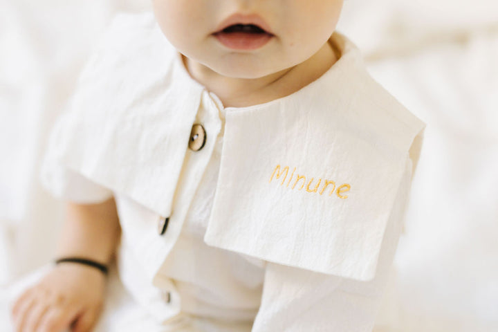 A close-up of a baby's face wearing a white outfit with a name tag.