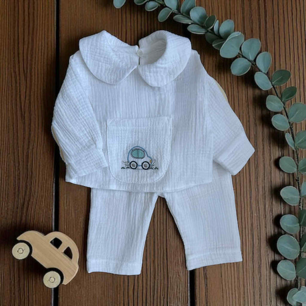 A white outfit with a small car embroidered on the front, placed on a wooden surface alongside a toy car and some greenery.