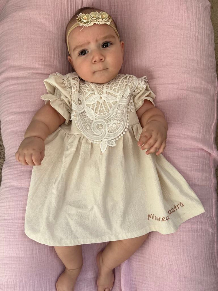 A baby girl wearing a white lace dress with a floral pattern lies on a pink blanket, looking directly at the camera.