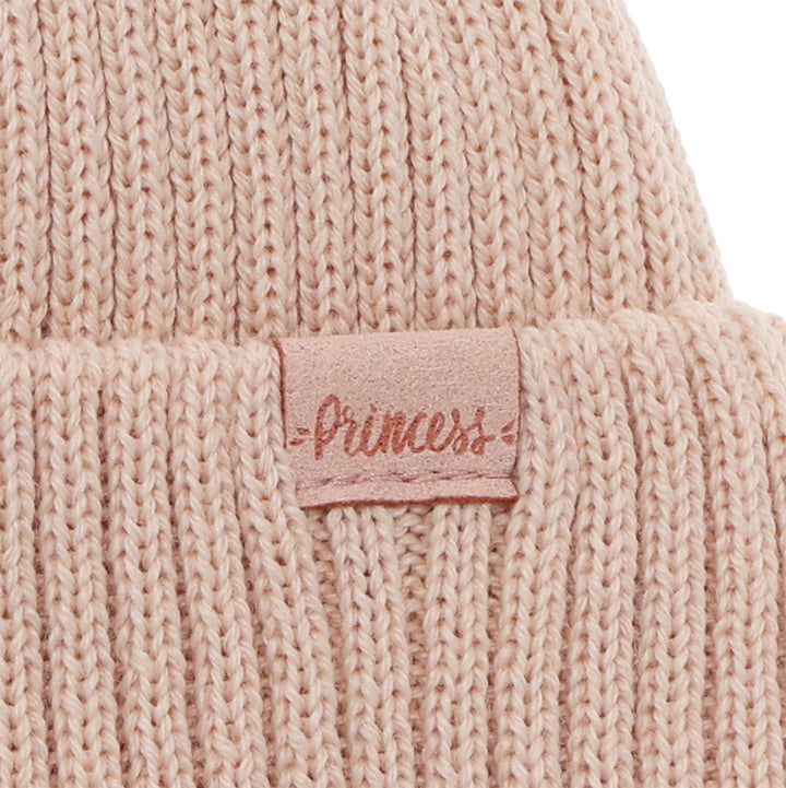 A close-up image of a beige knitted beanie with a small pink tag attached to it.