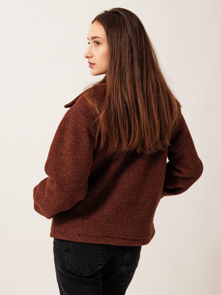 A woman with long brown hair wearing a brown sweater stands with her back to the camera, looking away from the camera.