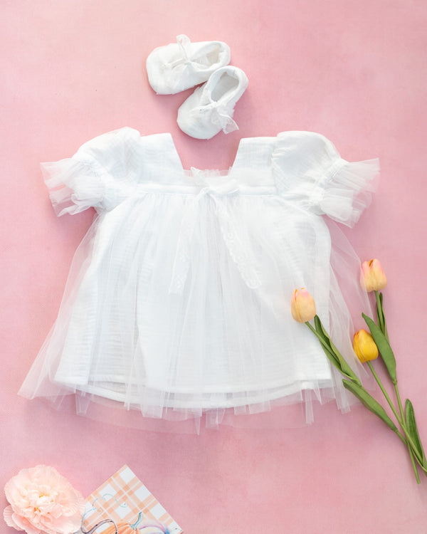 a white baby dress with a tulle skirt, a pair of white baby shoes, and a bouquet of yellow and pink tulips on a pink background.