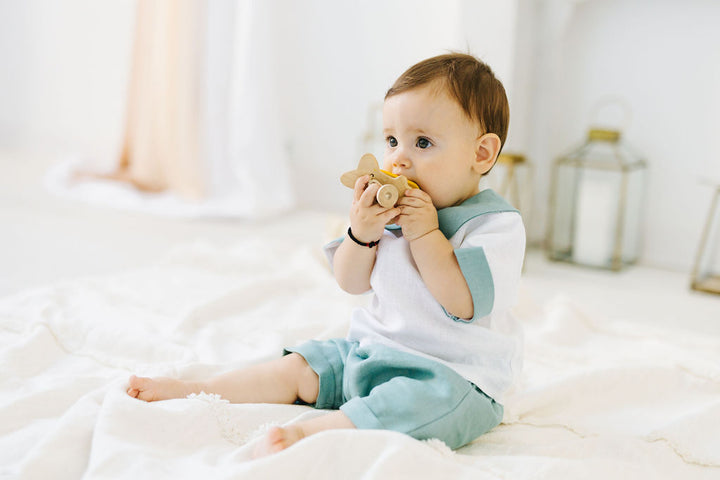 A baby boy sitting on a white blanket, holding a wooden toy in his mouth.