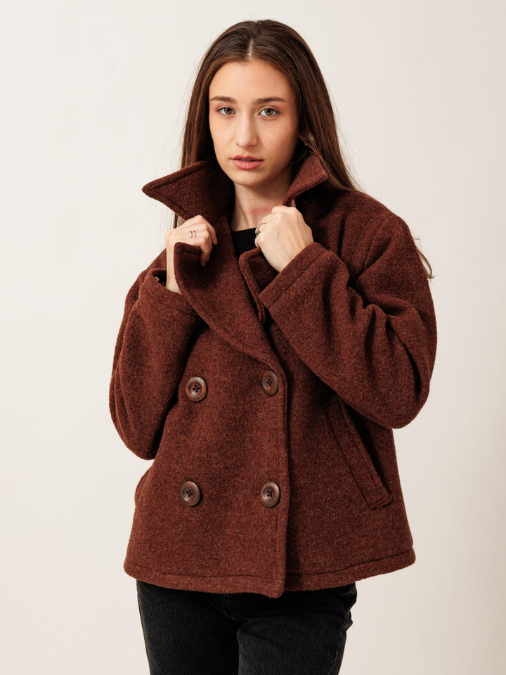 A young woman with long brown hair wearing a brown pea coat stands against a plain background, looking directly at the camera with a serious expression.