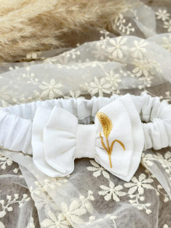 A white bow tie with a gold embroidered wheat stalk is displayed on a white lace background.