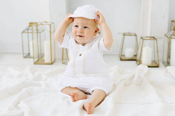 A smiling baby boy wearing a white outfit and hat sits on a white blanket, surrounded by gold lanterns.