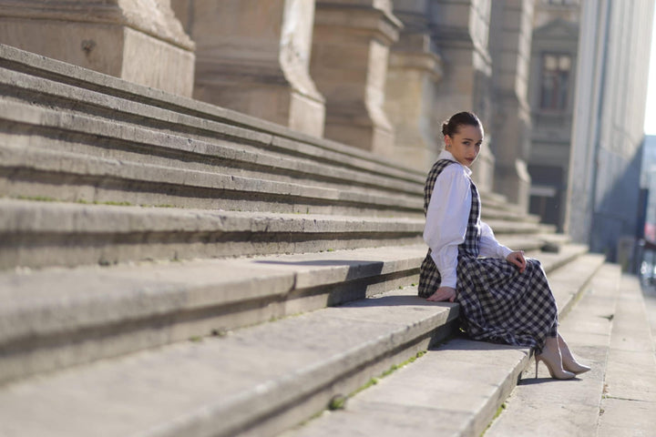A woman in a white blouse and black and white checkered dress sits on the steps of a building, looking away from the camera.