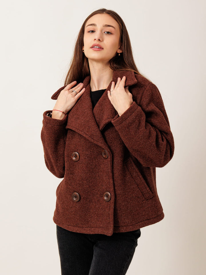 A young woman with long brown hair wearing a brown pea coat stands against a plain white background, looking directly at the camera with a neutral expression.