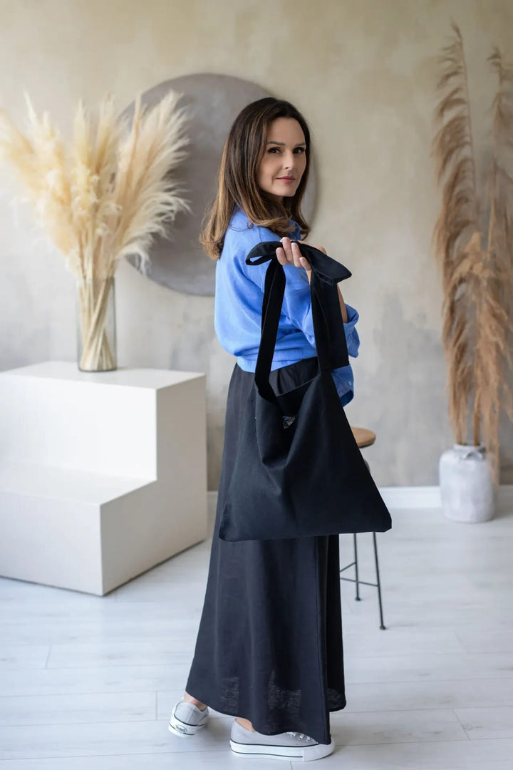 A woman in a blue shirt and black skirt stands in a minimalist room, holding a black bag and looking directly at the camera.