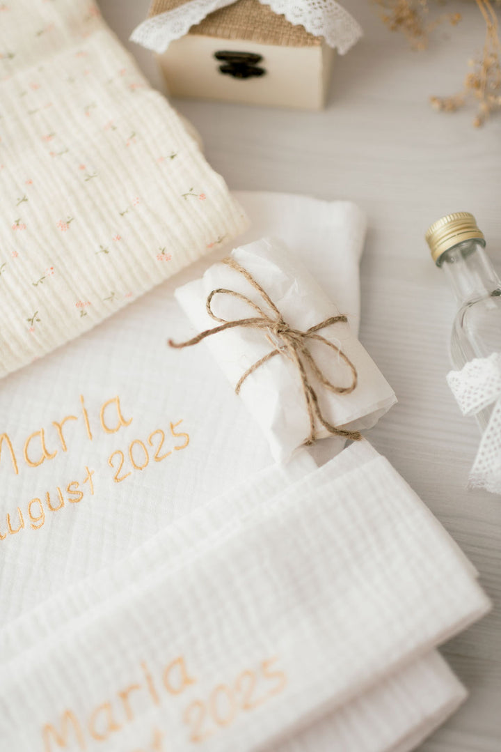 a close-up view of a white towel with the name "Maria" written on it, along with the date "August 25, 2015". The towel is tied with a brown ribbon, and there is a small box and a bottle of lotion on the table.