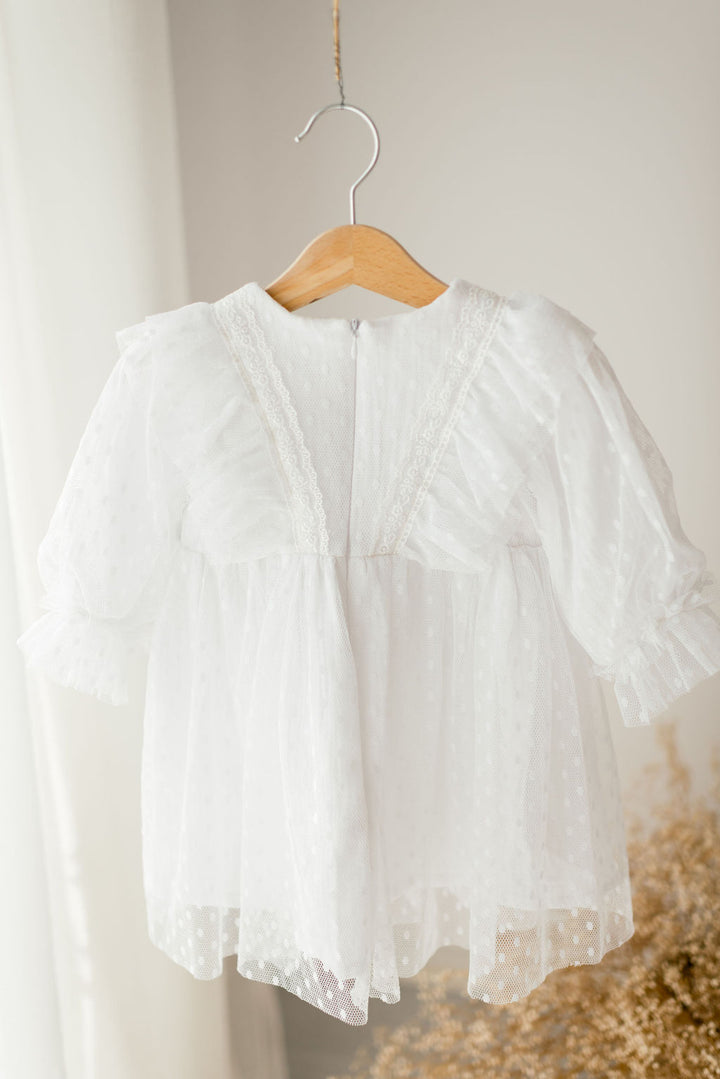 A white, ruffled dress with lace trim hangs on a wooden hanger against a white wall.