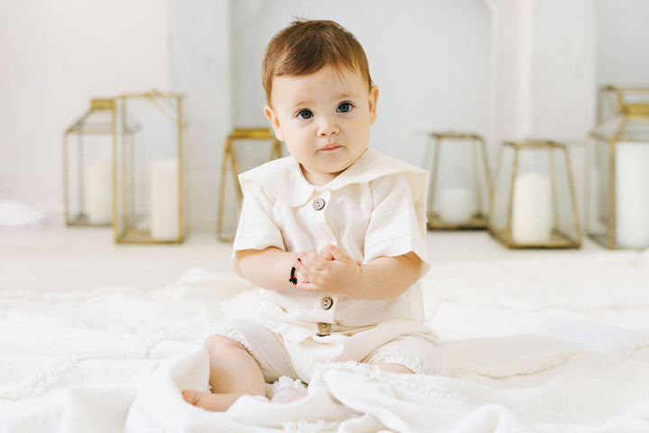 A baby in a white outfit sits on a white blanket, looking directly at the camera with a thoughtful expression.