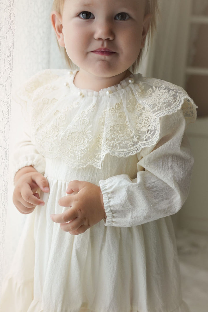 A young child in a white lace dress stands in front of a window, gazing directly at the camera with a thoughtful expression.