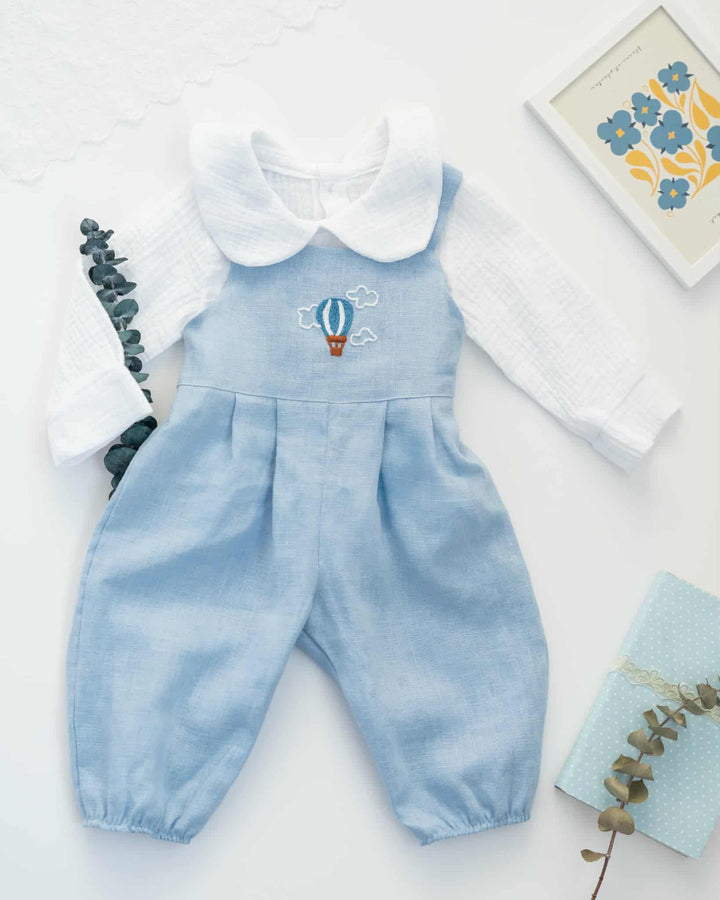 A light blue onesie with a white collar and a hot air balloon embroidered on the front, along with a white shirt, is displayed on a white background.