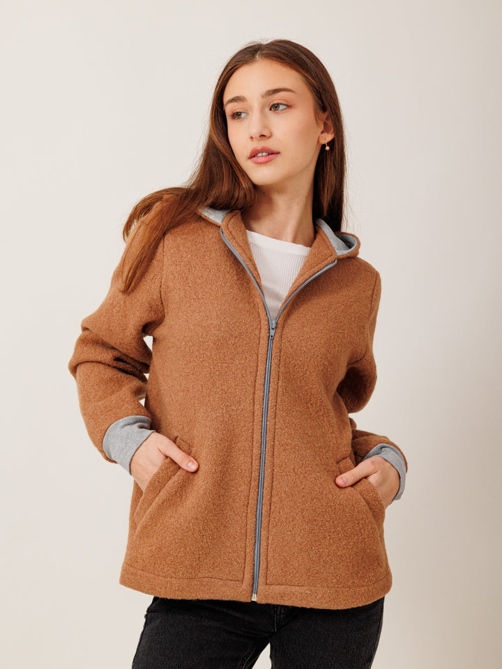 A young woman with long brown hair wearing a brown hooded jacket stands against a plain background.